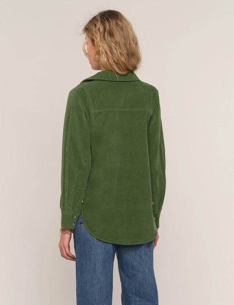 A woman viewed from the back wearing an olive green corduroy shirt jacket with blue jeans, set against a plain light beige background.