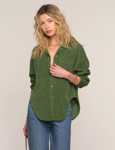A woman modeling an olive green corduroy button-up shirt with blue jeans, standing against a plain light beige studio background.
